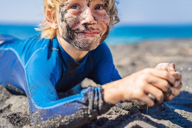 Black Friday Konzept lächelnder Junge mit schmutzigem schwarzem Gesicht sitzt und spielt am schwarzen Sandstrand