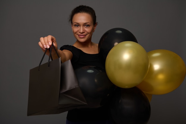 Black Friday-Konzept mit einer fröhlichen, attraktiven Frau mit schönem Lächeln, in Schwarz gekleidet, die schwarze und goldene Luftballons und Einkaufstasche hält und die Kamera im Hintergrund mit Kopien-Werberaum betrachtet