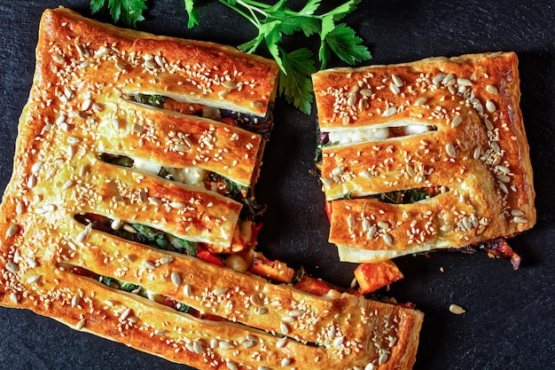 Blätterteig-Gemüsekuchen mit Ziegenkäse überbacken mit gerösteten Sonnenblumen- und Sesamsamen, gefüllt mit geröstetem Gemüse: Tomaten, Zwiebeln und Kürbis, auf schwarzem Schiefer, Draufsicht, Nahaufnahme