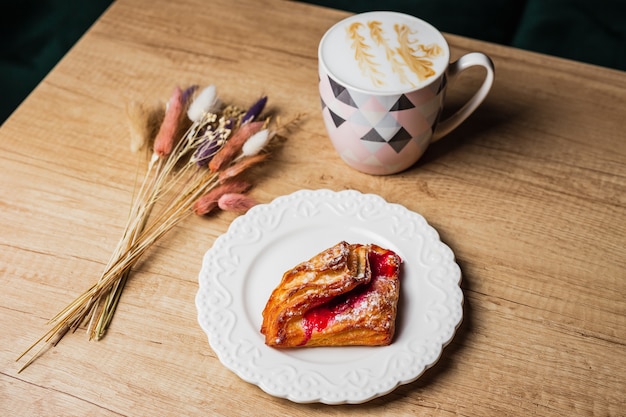 Blätterteig mit roter Marmelade auf einem weißen Teller, eine Tasse flachen weißen Kaffee und Blumen