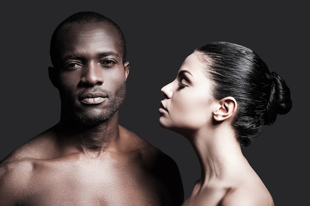 En blanco y negro. Retrato de un hombre africano sin camisa y una mujer caucásica que se unen entre sí mientras están de pie contra el fondo gris