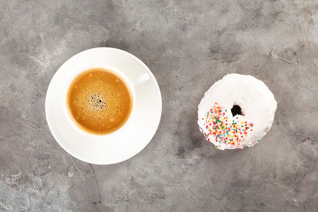 Blanco taza de café y donut en glaseado blanco y topping de pasteles de azúcar multicolor sobre fondo gris. Lay Flat