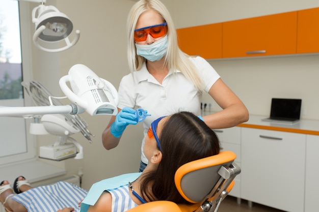 Blanqueamiento dental con dispositivo de blanqueamiento UV dental, asistente dental que atiende al paciente, ojos protegidos con gafas. Tratamiento de blanqueamiento con luz, láser, flúor. Blanqueamiento dental artificial.