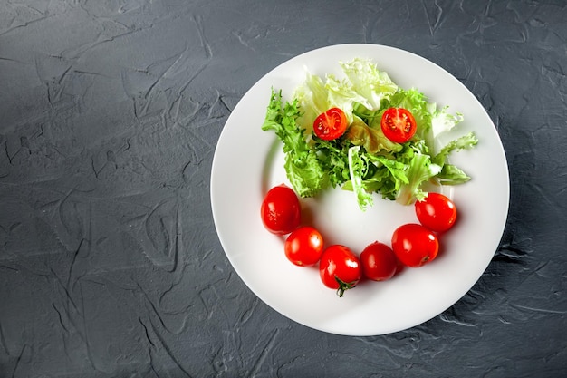 Blattsalat und Kirschtomaten in Form und Form eines Emoji auf einem weißen Teller auf grauem Hintergrund Draufsicht