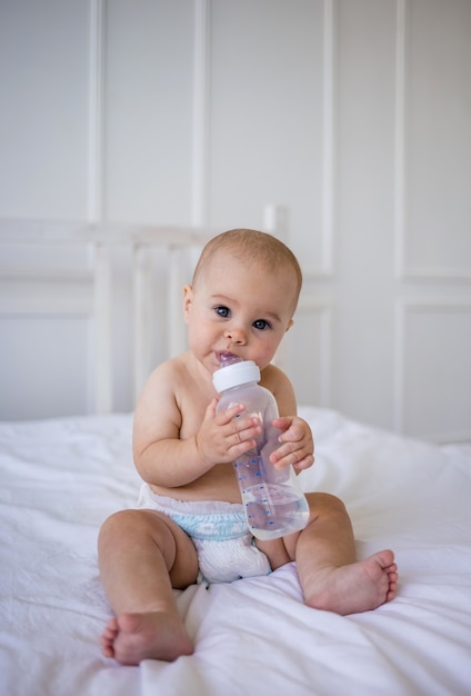 Blauäugiges Baby in einer Windel trinkt eine Flasche Wasser auf einer weißen Baumwolldecke auf dem Bett im Zimmer