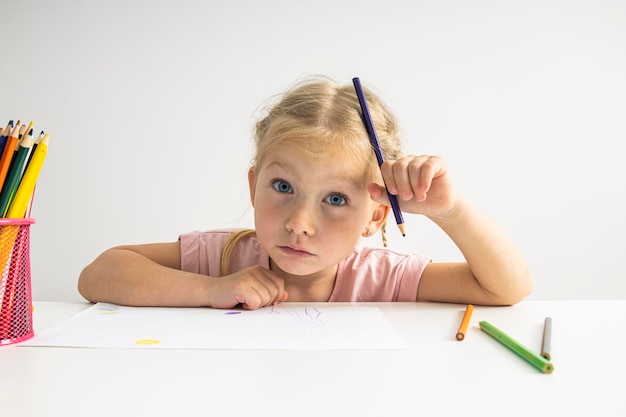 Blauäugiges blondes Mädchen zeichnet mit Buntstiften, die an einem weißen Tisch sitzen