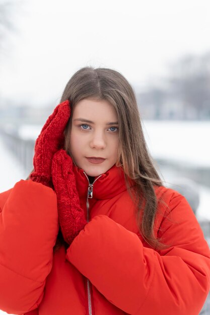 Blauäugiges schönes Mädchen in modischer roter Daunenjacke und gestrickten Fäustlingen posiert im Winter draußen