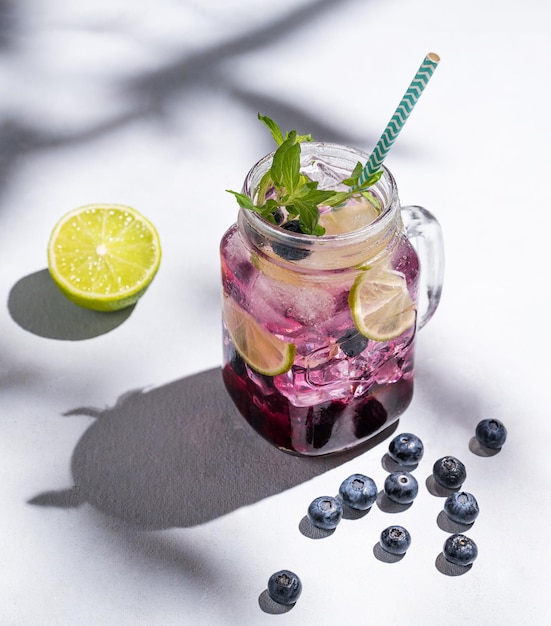 Blaubeer-Mojito oder Limonade mit Limetten-Eis und Minze in einem Glas auf einem hellen Hintergrund mit Beeren, Früchten und Schatten Das Konzept eines erfrischenden Sommergetränks