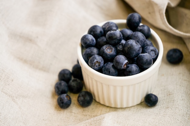 Blaubeerbeeren in einem weißen Tontopf mit verstreuten Beeren auf einem Leinenhandwerksstoff