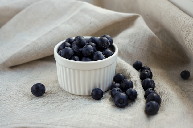 Blaubeerbeeren in einem weißen Tontopf mit verstreuten Beeren auf einem Leinenhandwerksstoff