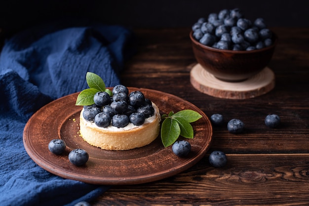 Blaubeerkuchen auf einem Tonteller mit Tektil auf dunklem Holzhintergrund, Sommerdessert, süßes Essen.