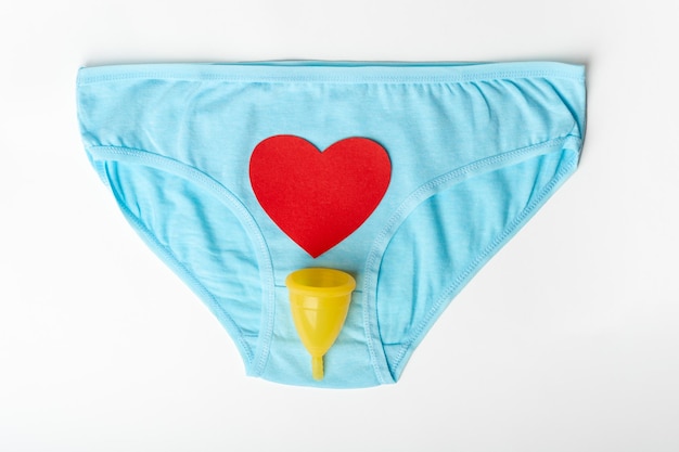 Blaue Damenunterwäsche und eine gelbe Menstruationstasse mit roter Herzform als Menstruationstropfen