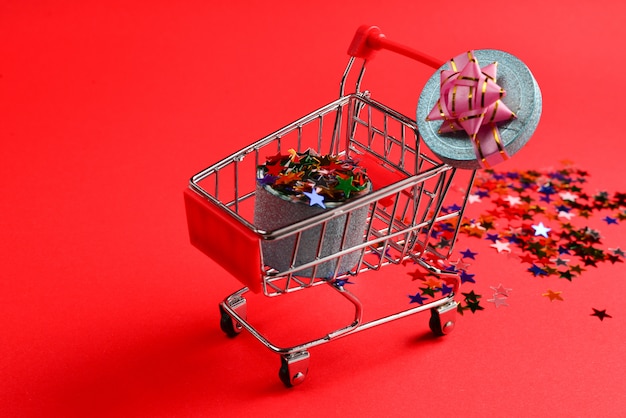 Blaue Geschenkbox mit rosa Schleife in einem Einkaufswagen und Konfetti auf einem roten Hintergrund.