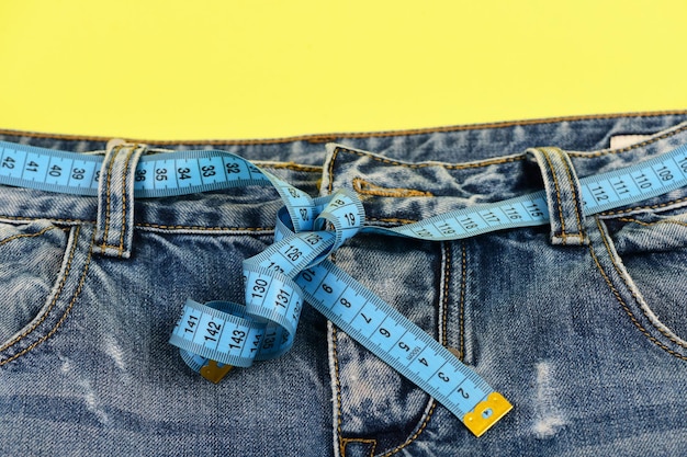 Blaue Jeans mit blauem Maßband statt Gürtel. Oberteil der Jeanshose isoliert auf gelbem Hintergrund. Gesunder Lebensstil und Diätkonzept. Nahaufnahme von Jeans mit Maßband um die Taille.