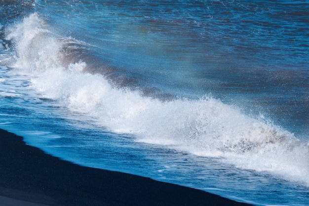 Blaue Meereswelle mit spritzenden Schlägen am schwarzen Sandstrand im Sommer