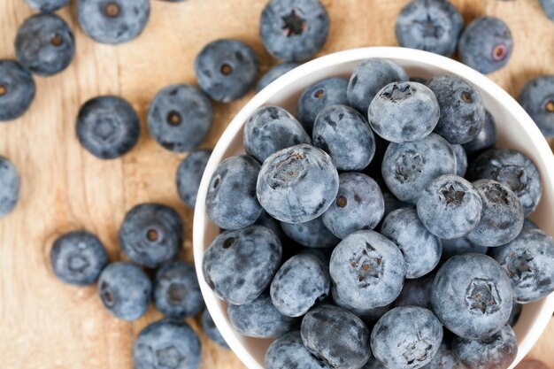 Blaue reife saftige Beeren Blaubeeren, die auf einer Holzoberfläche und einer runden Glasschale weiß liegen.