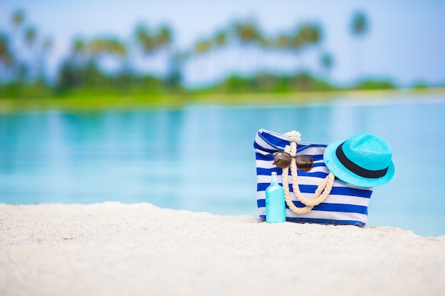 Blaue Tasche, Strohhut, Sonnenbrille und Sonnenschutzflasche auf weißem Strand