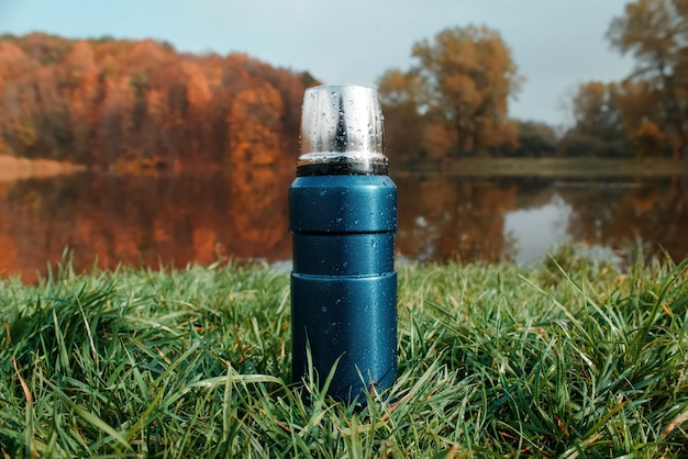 Blaue Vakuumthermosflasche auf grünem Gras, Herbstwald und See.