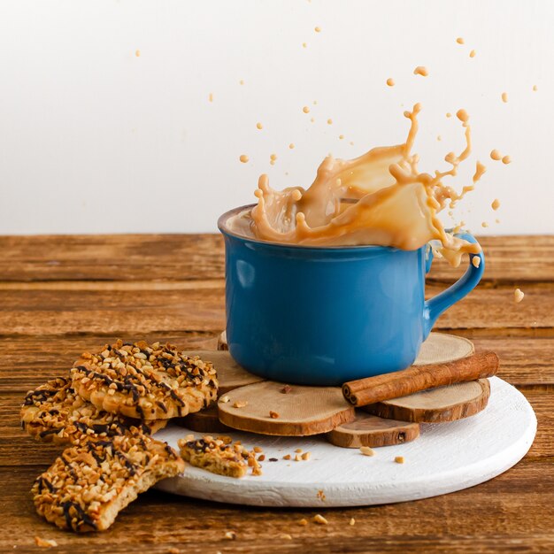 Blauer Becher mit dem Spritzen des Kaffees und der Plätzchen auf hölzernem Hintergrund.