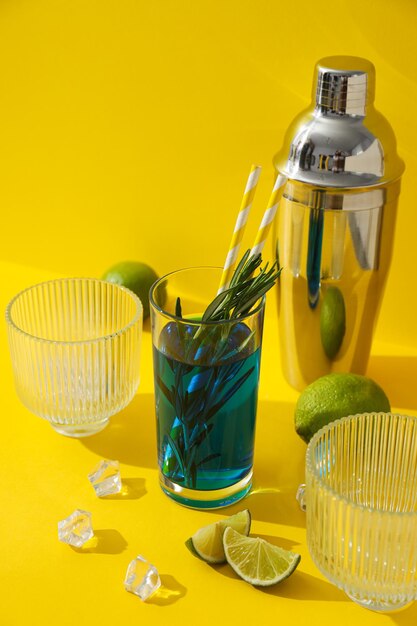 Blauer Cocktail in Glas, Limetten, Eiswürfel und Shaker auf gelbem Hintergrund