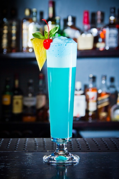 Blauer Getränkecocktail mit schaumdekorierter Ananas und Kirsche in großem Glas, nah an der Bartheke.