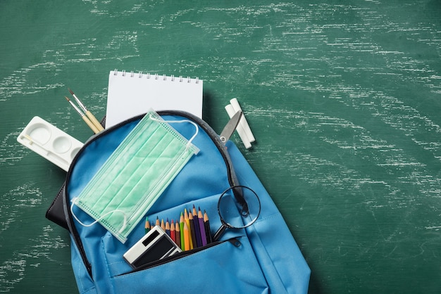 blauer Schulranzen-Rucksack an grüner Tafel mit Gesichtsmaskenschutz und Briefpapier