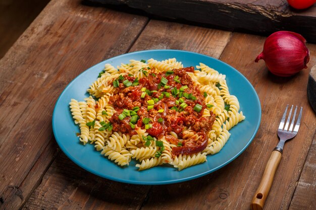 Blauer Teller mit Bolognese-Nudeln auf einem Holztisch neben einem Holzbrett-Gabelmesser und einer roten Zwiebel