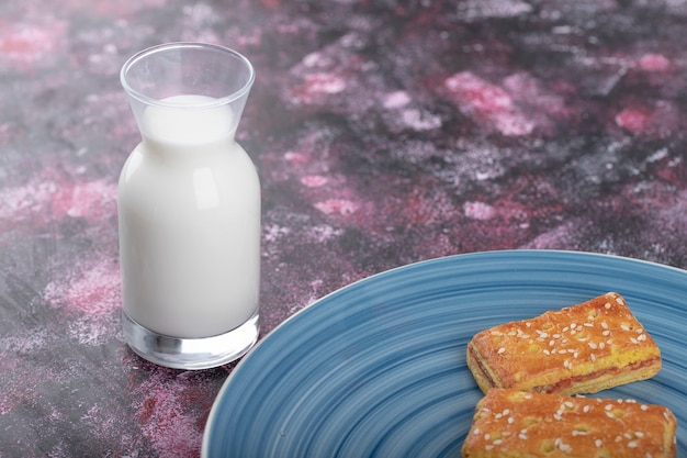 Blauer Teller mit Keksen mit Sesam und Glas Milch auf lila Tisch.