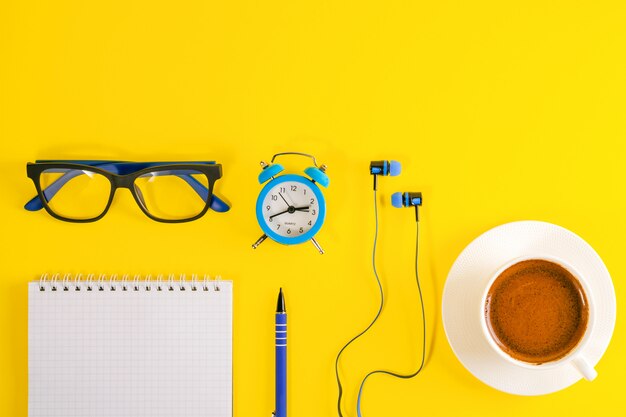 Blauer Wecker, Kopfhörer, Brille und Notizbuch mit Stift, auf gelbem Hintergrund. Kaffeetasse.