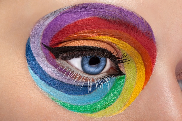 Blaues Auge mit künstlerischem Regenbogen-Make-up hautnah. Farben und bunt. Freude. Künstlerisches und modisches Make-up. Sucht machen. Kosmetika. Auf der Bühne schminken