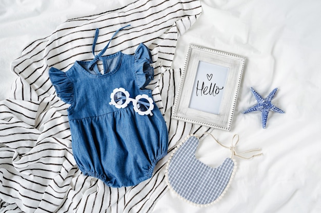 Blaues Babykleid mit Lätzchen, Sonnenbrille und Fotorahmen. Set Kinderkleidung und Accessoires für den Sommerurlaub auf dem Bett. Mode Neugeborenes. Flache Lage, Ansicht von oben