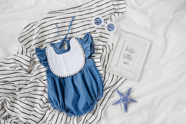 Blaues Babykleid mit Lätzchen, Sonnenbrille und Fotorahmen. Set Kinderkleidung und Accessoires für den Sommerurlaub auf dem Bett. Mode Neugeborenes. Flache Lage, Ansicht von oben