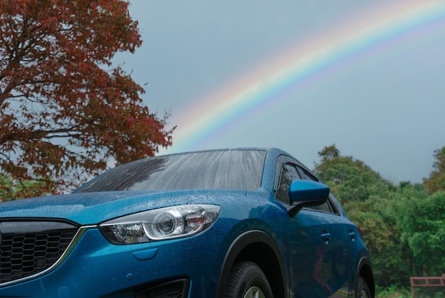 Blaues SUV-Auto, das mit Regentropfen gegen grünen Wald und Regenbogenhimmel bedeckt ist Auto im Park im Freien geparkt