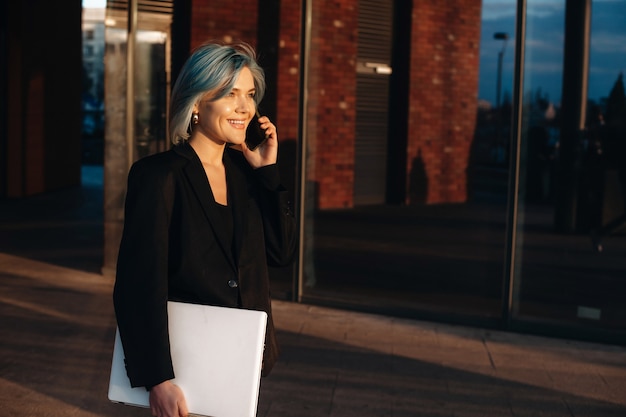 Blauhaariger kaukasischer Unternehmer, der eine Telefondiskussion beim Gehen mit einem Computer hat