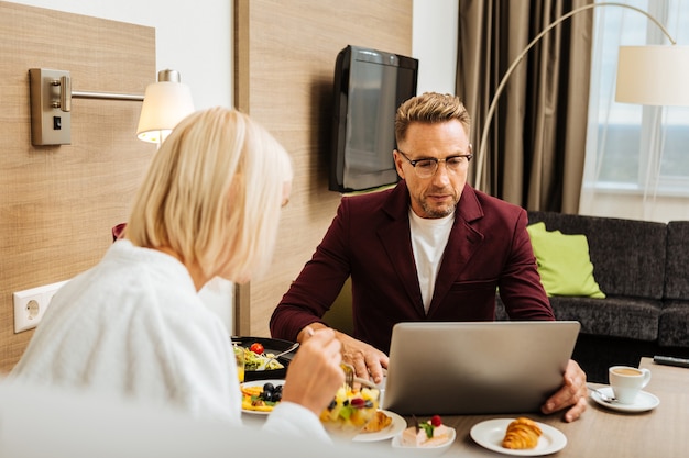 Blazer cor de vinho. Homem com corte de cabelo estiloso e óculos sentado com o laptop na mesa e sua amante contra ele