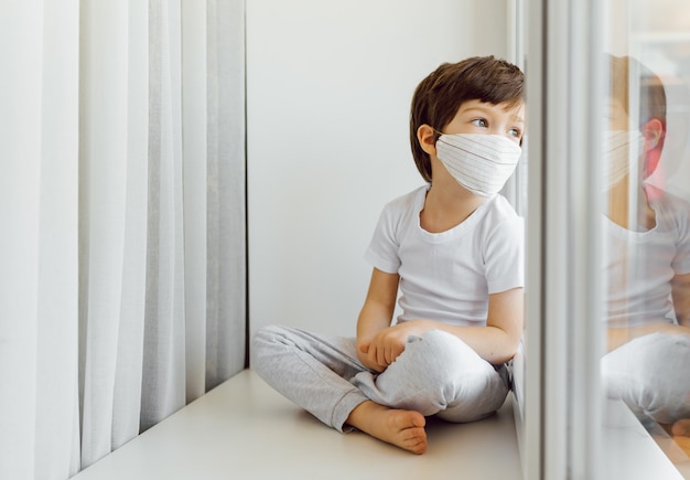 Bleiben Sie zu Hause Quarantäne Coronavirus Pandemie Prävention. Trauriges Kind sowohl in medizinischen Schutzmasken in der Nähe von Fenstern als auch aus dem Fenster