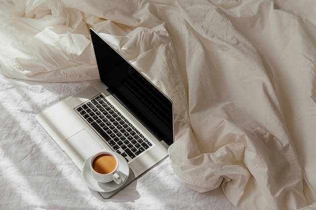 Bleiben Sie zu Hause, Quarantäne. Von zu Hause aus arbeiten. Laptop und Tasse Kaffee auf weißem Bett mit einer Decke. Konzept zu Hause arbeiten. Morgenlicht