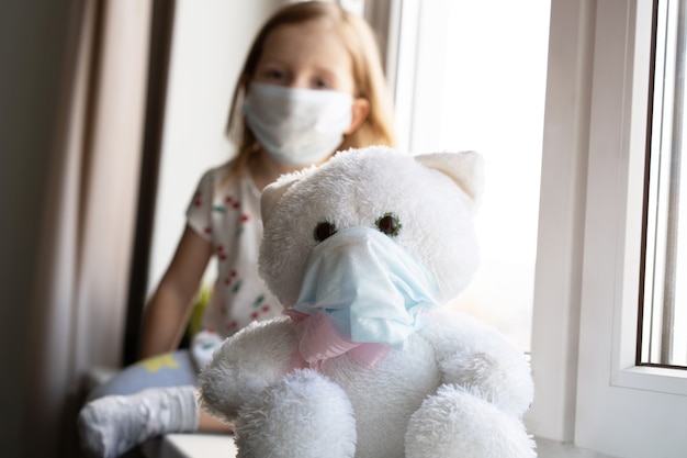 Bleiben Sie zu Hause unter Quarantäne zur Vorbeugung einer Coronavirus-Pandemie. Trauriges Kind und sein Teddybär sitzen beide in medizinischen Schutzmasken auf der Fensterbank und schauen aus dem Fenster. COVID-2019-Krankheitskonzept