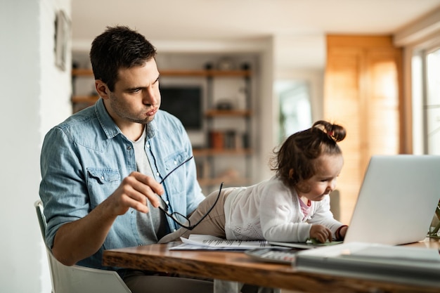 Bleiben Sie zu Hause Vater, der sich gestresst fühlt, während er babysittet und versucht zu arbeiten, während seine kleine Tochter seinen Laptop benutzt