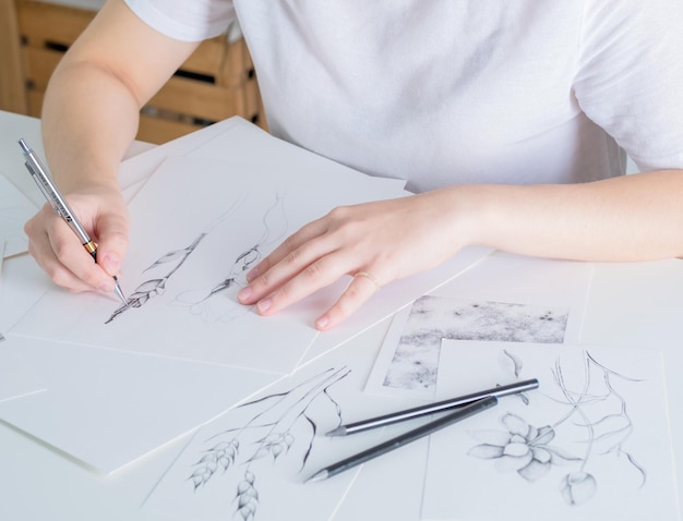 Bleistiftzeichnungen liegen auf dem Tisch Eine junge Frau mit europäischem Aussehen zeichnet mit einem Bleistift auf weißem Papier Grafiken Der Prozess des Zeichnens mit einem Bleistift zu Hause an einem weißen Tisch