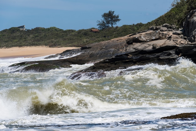 Blick auf den strand von areia negras in rio das ostras in rio de janeiro mit sonnigem tag, blauem himmel und einigen wolken. starkes meer und schwarzer sand gemischt mit gelb und vielen felsen.