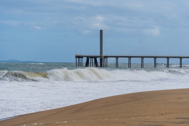 Blick auf den Strand von Rio das Ostras in Rio de Janeiro mit dem Pier des Abgesandten. Sonniger Tag, blauer Himmel und einige Wolken. Starkes Meer und gelblicher Sand und viele Felsen.