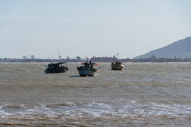 Blick auf den Strand von Rio das Ostras mit dem Zusammenfluss des Flusses in Rio de Janeiro. Sonniger Tag, blauer Himmel. Gelber Sand und einige Felsen. Holzbrücke zu überqueren. Boote und Angeln am Pier.