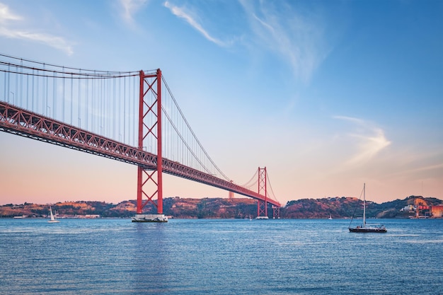 Blick auf die Brücke 25 de Abril über den Fluss Tejo, das Christus-König-Denkmal und ein Yachtboot bei Sonnenuntergang in Lissabon, Portugal