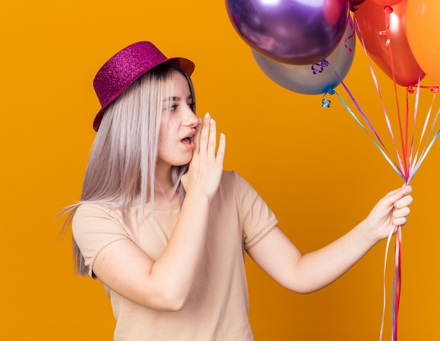 Blick auf die Seite junges schönes Mädchen mit Partyhut mit Luftballons flüstert isoliert auf oranger Wand