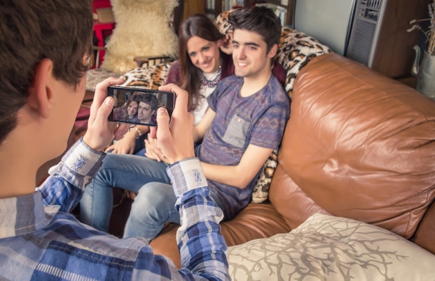 Blick auf einen jungen Freund, der Fotos mit einem Smartphone macht, um Hipster-Teenager-Paar auf dem Sofa zu sitzen. Konzentrieren Sie sich auf den Telefonbildschirm.