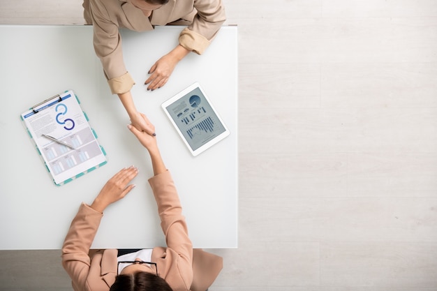 Blick auf zwei junge zeitgenössische Geschäftsfrauen, die den Schreibtisch über den Tisch schütteln, nachdem sie einen Deal zwischen Finanzinformationen in Tablet und auf Papier gemacht haben