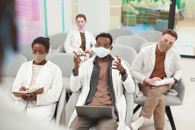 Blick aus der Vogelperspektive auf eine multiethnische Gruppe von Menschen, die Masken und Laborkittel tragen, während sie Vorlesungen über Medizin im College oder Coworking Center hören, Platz kopieren