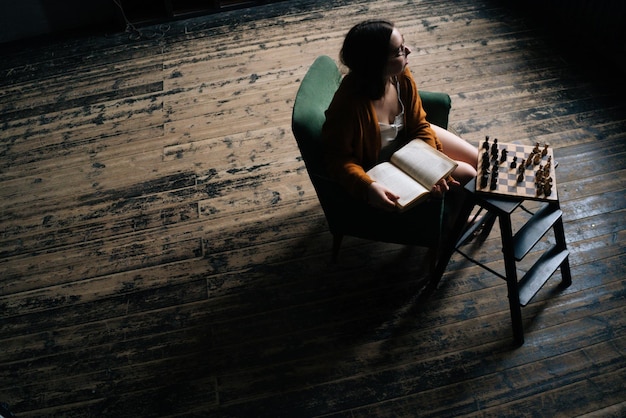 Blick aus der Vogelperspektive auf eine nachdenkliche junge Frau mit eleganter Brille, die ein Buch hält, das in einem Sessel in einem dunklen Vintage-Raum sitzt und wegschaut