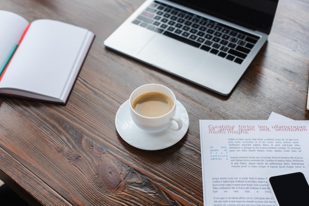 Blick aus der Vogelperspektive auf eine Tasse Kaffee in der Nähe von Copybook-Laptop und Smartphone auf Papieren auf dem Tisch im Büro